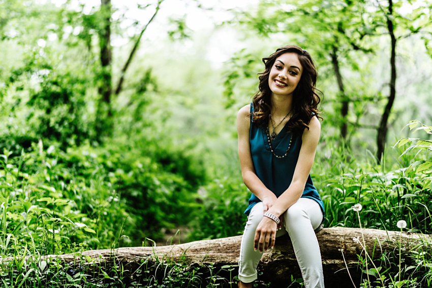 girl in white pants and a blue top sitting on a log in the forest