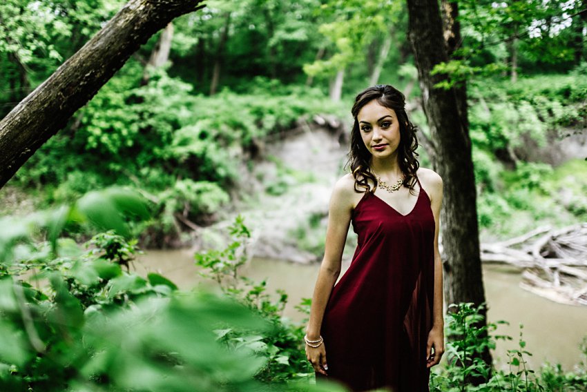 girl in a red dress standing in the woods in front of a river looking at the camera