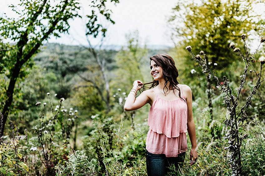 girl in a pink top standing on a tall hill next to a forest in the fall while playing with her hair