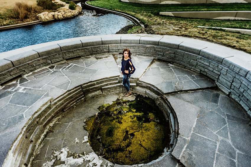 girl in a blue top and jeans standing in a spiral brick structure that leads to a pool of mossy water