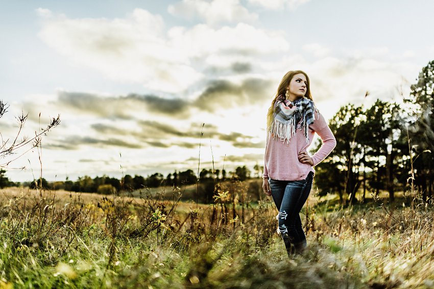 girl in a pink sweater and scarf standing in the prairie grass with a sunset behind her