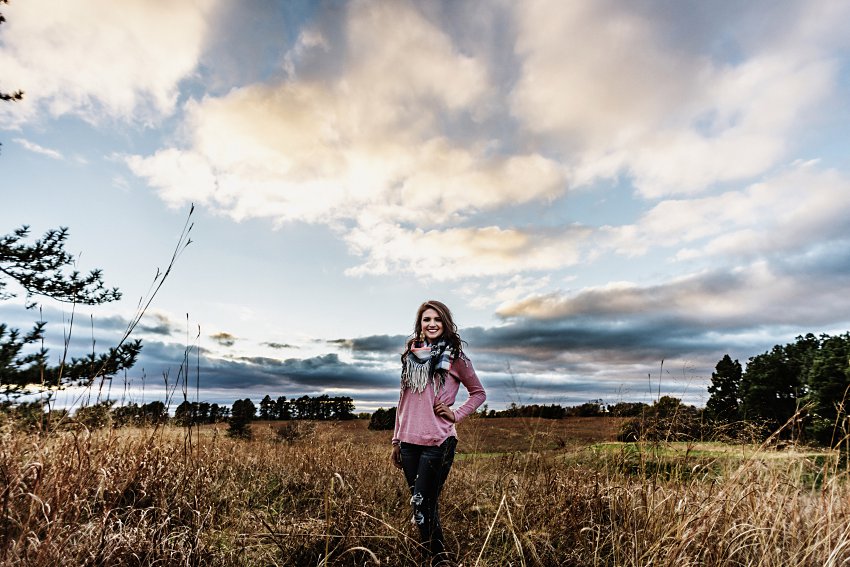 girl in a pink sweater and scarf standing in the prairie with lots of clouds overhead