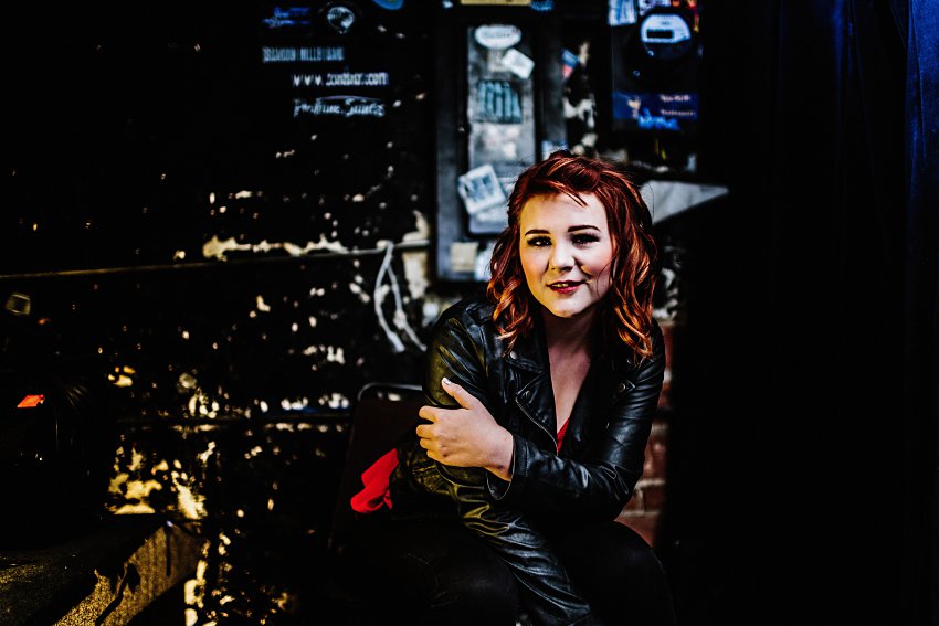girl in red top and leather jacket sitting in front of an old wall