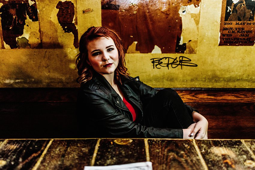 girl in red top and leather jacket sitting in an old booth against the wall with torn posters and graffiti behind her