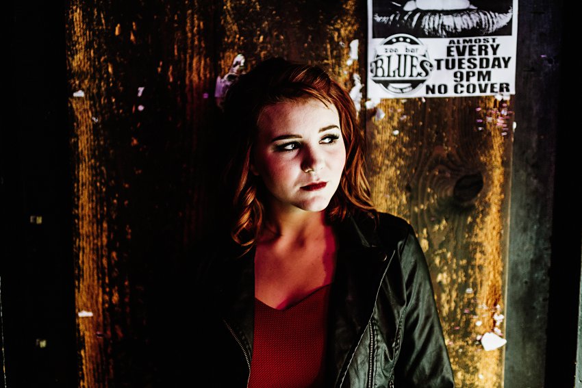 girl in red top and leather jacket standing in front of an old wall with band posters behind her
