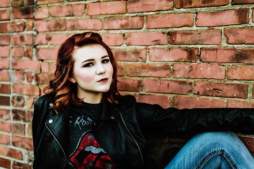 girl in red top and leather jacket sitting in an alley in front of a brick wall