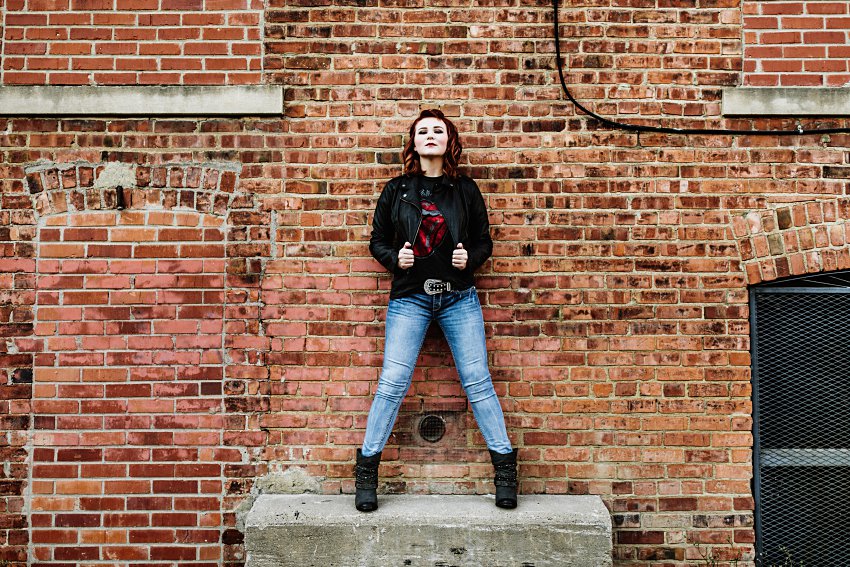 girl in red top and leather jacket standing on a concrete block in an alley in front of a brick wall