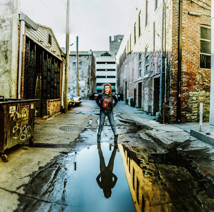 girl in red top and leather jacket standing in an alley in front of her reflection in a pool of water