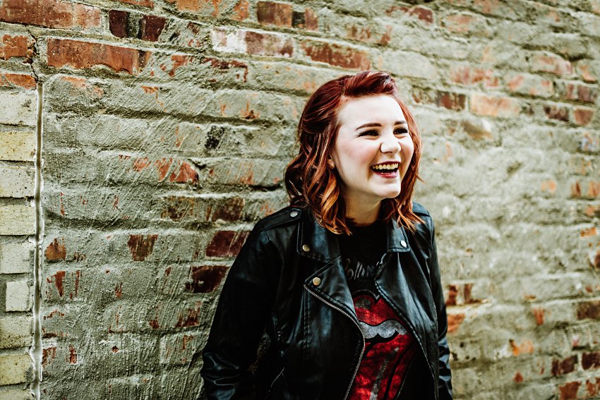 girl in red top and leather jacket in front of a brick wall laughing