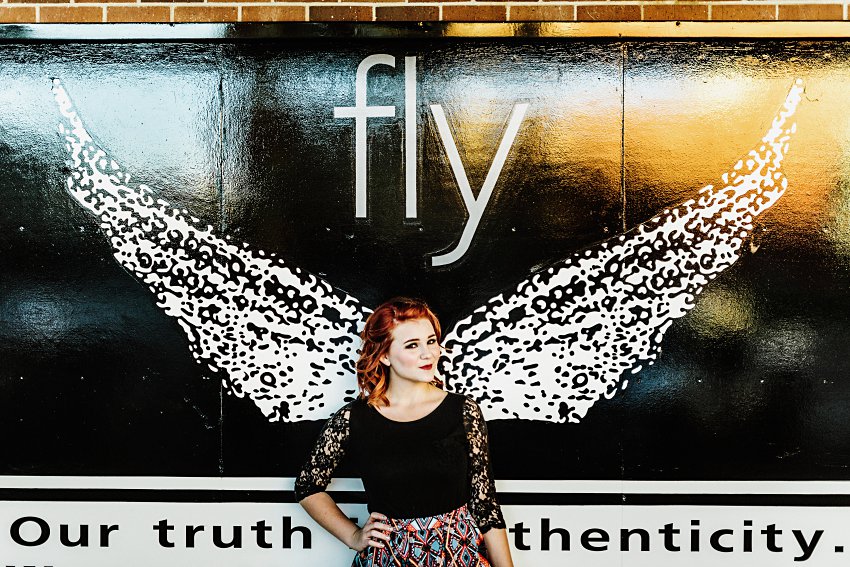 girl in a black top and colorful patterned skirt standing in front of a big poster with wings and the word fly