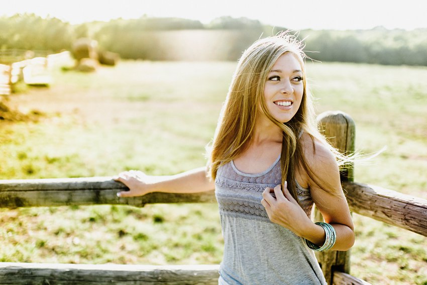 girl in a gray tank top standing by a fence and open field in sunset