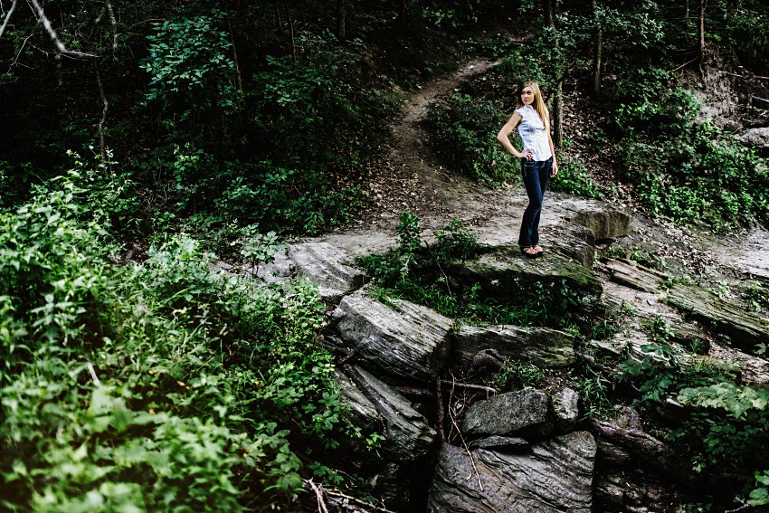 girl in a white top and black pants standing on a rocky ledge in the woods