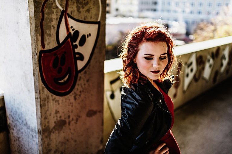 close up of a girl in red top and leather jacket looking down standing in a parking garage with music graffiti in the background